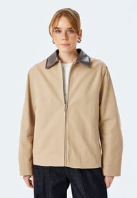 Beige jakke med brunt læderkrave, lynlås foran og lange ærmer. Lavet af struktureret stof, med sidelommer og en afslappet pasform.