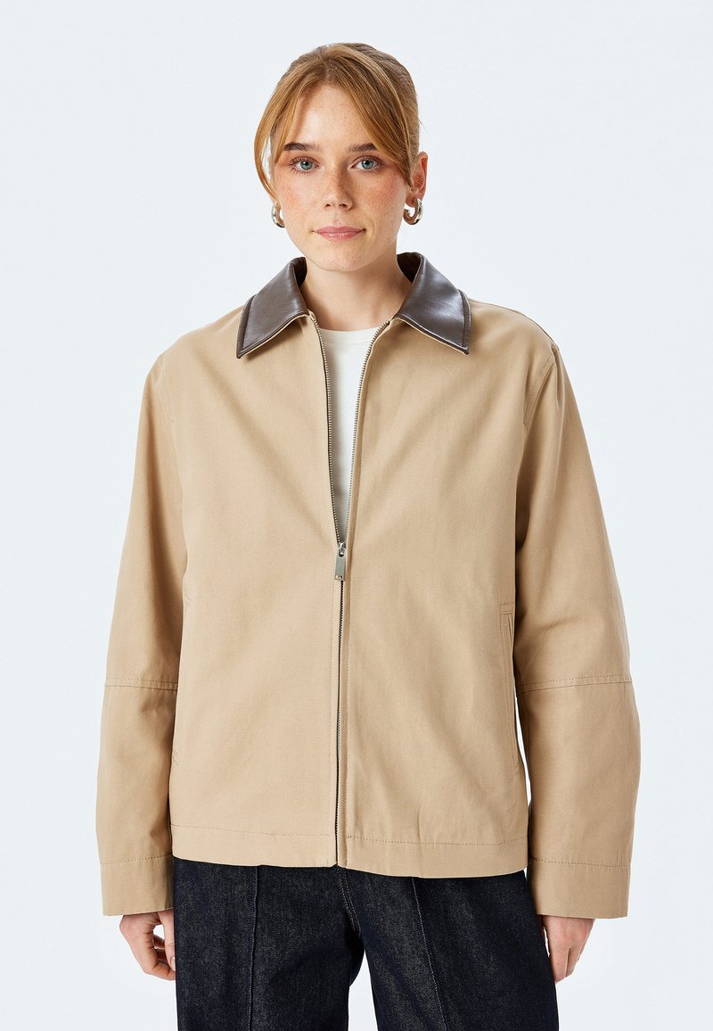 Beige jakke med brunt læderkrave, lynlås foran og lange ærmer. Lavet af struktureret stof, med sidelommer og en afslappet pasform.