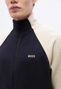 Μπουφάν με φερμουάρ σε ναυτικό μπλε και κρεμ από υφή υφάσματος, με ψηλό γιακά και κεντημένο λογότυπο "BOSS" σε μπεζ στο στήθος.