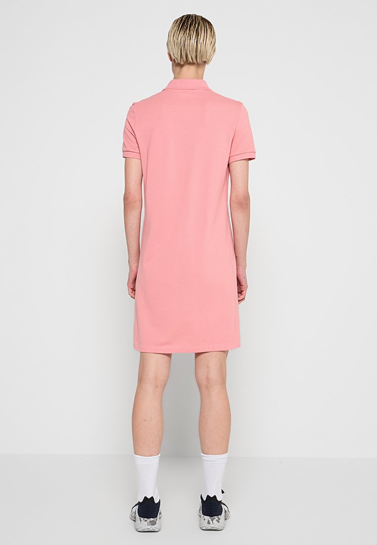 Robe polo rose en matériau doux, dotée d'un col, de manches courtes et d'un ourlet droit, portée avec des chaussettes blanches et des baskets grises.
