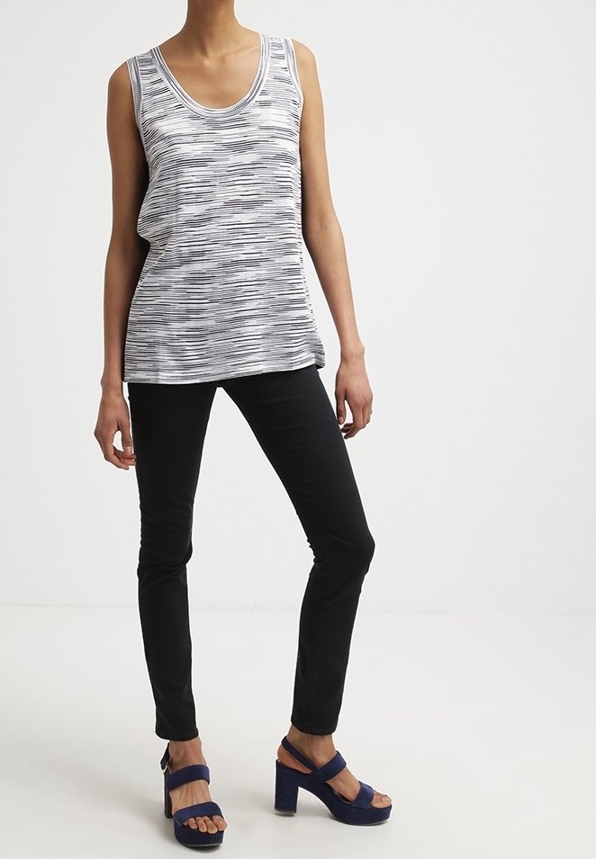 Femme portant un haut sans manches rayé blanc et noir, un pantalon noir ajusté et des sandales à plateforme bleu marine sur fond blanc.