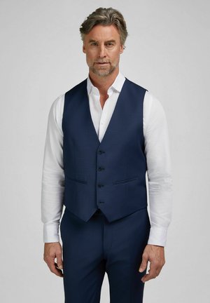 Homme aux cheveux poivre et sel portant une chemise blanche, un gilet bleu marine à trois boutons et un pantalon assorti, debout devant un fond uni.
