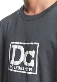 Dunkelgraues Baumwoll-T-Shirt mit einem weißen Grafiklogo und dem Text "DC" sowie "DCSHOES - 1994". Klassischer Rundhals-Design, weicher Stoff.