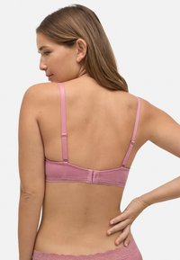 Sujetador rosa con tirantes ajustables, tejido suave, cierre de gancho y ojal, y adornos de encaje en la banda. La vista trasera muestra la simplicidad del diseño.