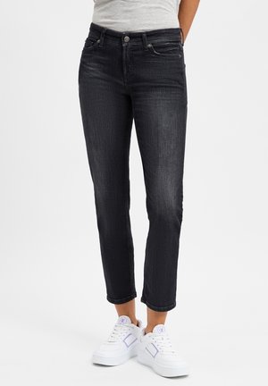 Femme portant un jean noir slim coupé au-dessus des chevilles et des baskets blanches avec un logo violet, debout les mains derrière le dos.