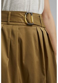 Khaki nederdel med plisseringer, der har en bred talje og en rektangulær skildpaddeagtig spænde. Stoffet har en glat, let skinnende tekstur.