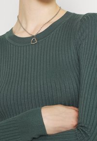 Ribbed vihreä pitkähihaine paita, jossa on pyöreä kaula-aukko. Sisältää hopeaketjun kaulaketjun, jossa on sydämen muotoinen riipus. Teksturoitu kangas näkyy yksityiskohtana.