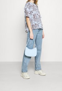 Blå grafisk t-shirt med bruna mönster, kombinerad med en ljusblå handväska och ljusa denimjeans. Modellen bär ljusa sneakers.