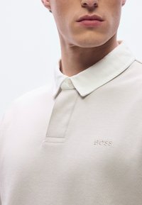 Tricou polo bej deschis cu guler alb, prezentând o fâșie cu trei nasturi și logo-ul brodat „BOSS” pe piept. Textură netedă.