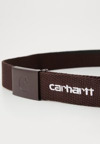 Bruine geweven stofriem met een rechthoekige bruine gesp, voorzien van het witte "Carhartt" logo en een gladde textuur.
