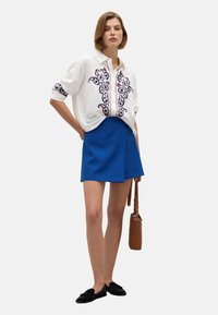 Camicia bianca con ricami paisley blu e rossi, abbinata a una gonna avvolgente blu. Modella tiene una borsa marrone e indossa ballerine nere.