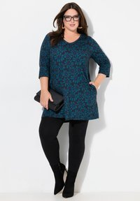 Türkisfarbener Tunika mit floralem Muster, Dreiviertelärmel, kombiniert mit schwarzen Leggings und Stiefeletten. Hält eine schwarze Clutch.