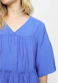 Top leggero blu con scollatura a V, maniche corte e vita arricciata. Il tessuto ha una finitura leggermente testurizzata e una vestibilità rilassata.