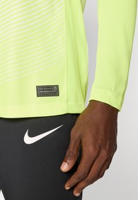 T-shirt de sport à manches longues de couleur jaune fluo avec des rayures blanches diagonales ; comporte une étiquette noire avec du texte. Associé à un pantalon noir portant un logo blanc.