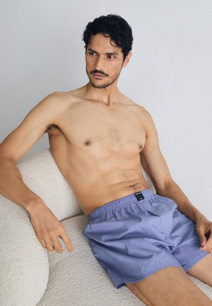 Shorts en coton violet clair avec une taille élastique, arrivant au-dessus du genou, portés par un modèle assis sur un fond neutre.