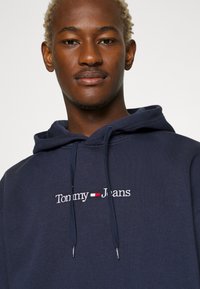 Sudadera azul marino hecha de una mezcla de algodón con capucha ajustable; cuenta con un logo bordado de "Tommy Jeans" en la parte delantera.