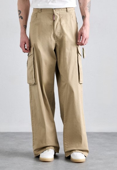 Pantaloni cargo beige con design a gamba larga, dotati di tasche laterali e una texture liscia. Abbinati a sneakers bianche.