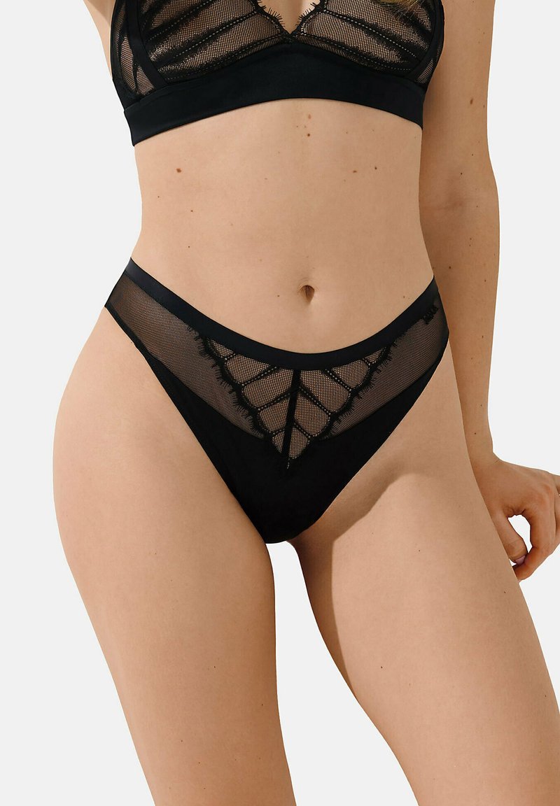Zwart kanten bralette en bijpassende thong met doorzichtige meshpanelen, voorzien van een geometrisch patroon en subtiele scalloped randen. Zacht, rekbaar materiaal.