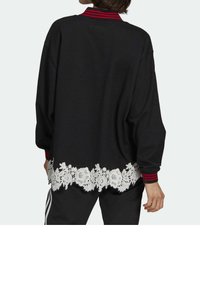 Mustat ylisuuret collegepaidat, joissa on valkoinen pitsireunus helmakohdassa ja punaraidallinen kaulus. Pitkät hihat, rento muotoilu, pehmeä tekstuuri.