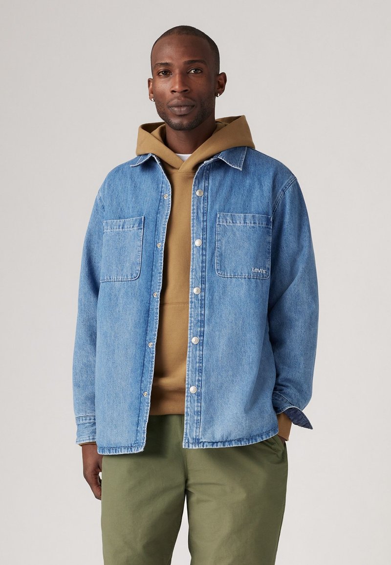 Camisa de mezclilla azul claro con dos bolsillos frontales, diseño de botones, usada sobre una sudadera marrón, combinada con pantalones verdes oliva.
