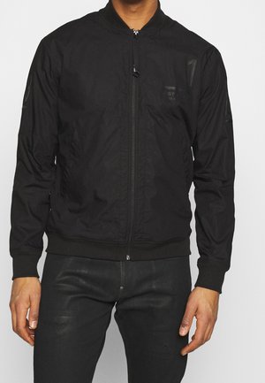 Mand iført sort bomberjakke med lynlås og sorte bukser, stående med armene afslappet ned langs siderne mod en ensfarvet baggrund.