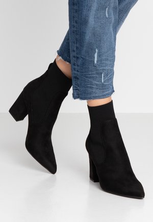 Black suede ankle boots with chunky heels worn with cropped distressed blue jeans on white floor.