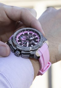 Montre-bracelet avec un cadran noir et rose, des accents argentés, un grand boîtier rond et un bracelet en caoutchouc rose. Elle possède des sous-cadrans chronographe et un indicateur de date.
