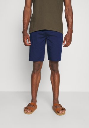 Mann trägt dunkelblaue Shorts, braune Sandalen mit Schnallen und ein olivgrünes Hemd, steht vor einem schlichten hellen Hintergrund.