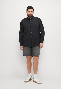 Schwarzes langärmliges Hemd mit Stehkragen, kombiniert mit grauen Jeansshorts und weißen Sneakers mit schwarzen Akzenten.