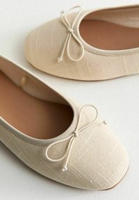 Bailarinas beige hechas de tela texturizada, con punta redondeada, suela plana y un lazo decorativo en la parte frontal.