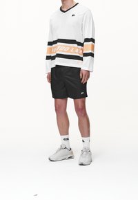 Camicetta a maniche lunghe in rete bianca con strisce nere e arancioni e testo, abbinata a pantaloni corti neri e sneakers grigie. Sfondo bianco uniforme.