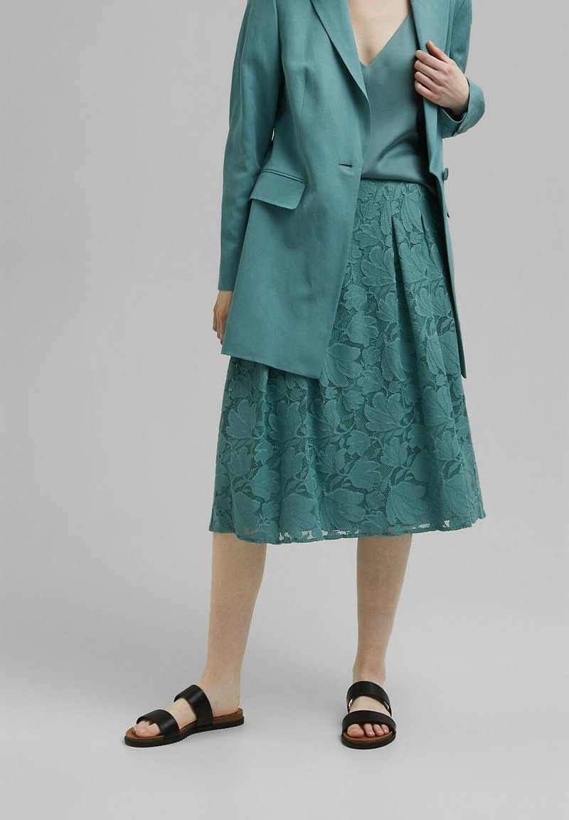 Blazer verde azulado y falda de encaje con un patrón floral, con un diseño plisado y textura en capas. Combinados con sandalias negras tipo slide.