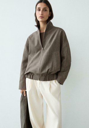 Femme portant une veste marron à fermeture éclair avec taille élastique et pantalon blanc, tenant un vêtement marron assorti devant un fond clair uni.