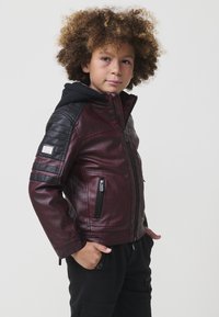 Ragazzo con capelli ricci che indossa una giacca di pelle bordeaux e nera con tasche con cerniera, in piedi con le mani nelle tasche dei pantaloni neri, rivolto di lato.