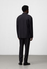 Completo nero a righe sottili con giacca a collo di taglio ampio e pantaloni con gamba dritta. Il tessuto sembra strutturato, abbinato a scarpe basse.