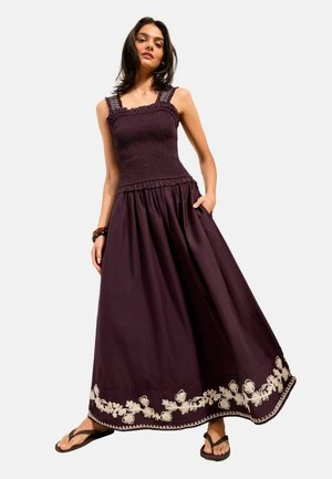 Žena má na sebe tmavofialové maxišaty bez rukávov s kvetinovou výšivkou pri leme, v kombinácii s čiernymi sandálmi a náramkom.