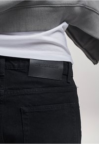 Schwarze Jeans aus Denim mit glatter Textur, ausgestattet mit einem Lederetikett mit der Aufschrift "2Y STUDIOS" am Bund, kombiniert mit einem grauen Sweatshirt.