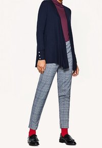 Marineblauer Cardigan mit vorderem Drapé und geknöpften Manschetten über einem burgunderfarbenen Rollkragenpullover, kombiniert mit karierten Hosen und schwarzen Schuhen, die mit roten Socken ergänzt werden.