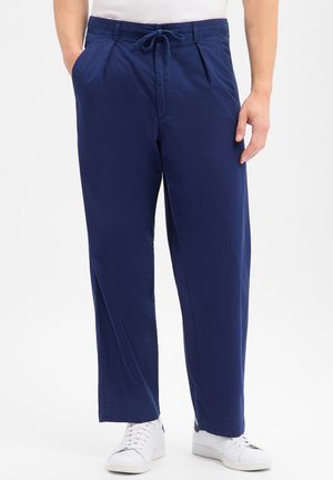 Homme portant un pantalon bleu marine à cordon et des baskets blanches, debout avec une main dans la poche devant un fond uni.