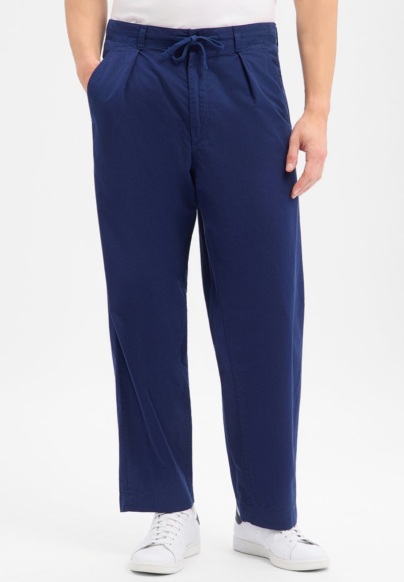 Homme portant un pantalon bleu marine à cordon et des baskets blanches, debout avec une main dans la poche devant un fond uni.