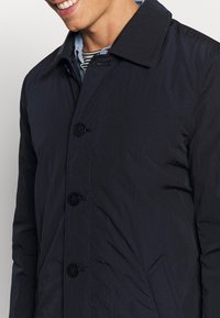 Marineblauer Steppmantel mit strukturierter Oberfläche, Reverskragen und vier schwarzen Knöpfen. Mit Seitentaschen und minimalistischem Design.