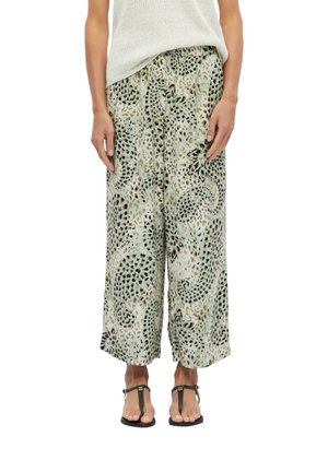 Femme portant un pantalon ample à jambes larges avec un motif animalier, un haut blanc sans manches et des sandales noires, debout, mains détendues.