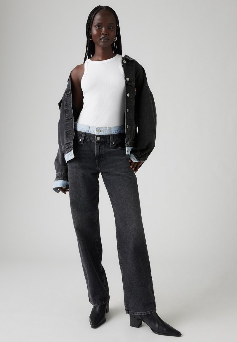 Veste en denim noir sur un débardeur blanc, associée à un jean noir taille haute et des bottines à talons noires. Design simple avec des accents minimaux.