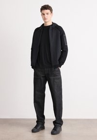 Črna zip-up jakna s kapuco, v kombinaciji s črnim majico, temno modrimi jeans hlačami in temnimi supergami. Jakna ima stranski žep.