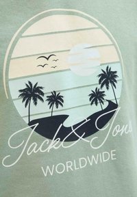 Camiseta verde menta con un gráfico circular que presenta palmeras, una puesta de sol y pájaros. El texto dice "Jack & Jones" y "WORLDWIDE" en blanco.