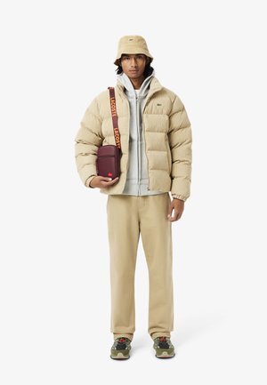 Beige puffer jacket, grey hoodie, cream pants, green bucket hat, maroon crossbody bag with Lacoste logo, and green sneakers with orange accents.
