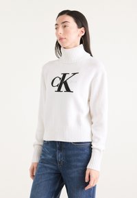 Hvit strikket genser med høy ribbestrikket krage, med en stor svart "CK" logo foran. Bært med blå jeans, ribbestrikkede mansjetter og kant.