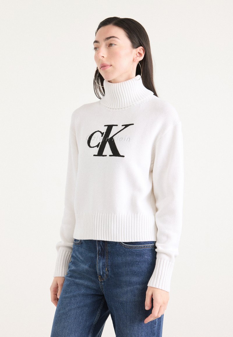Hvit strikket genser med høy ribbestrikket krage, med en stor svart "CK" logo foran. Bært med blå jeans, ribbestrikkede mansjetter og kant.