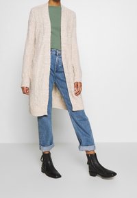 Stickad lång cardigan i ljusbeige med öppen front, texturerad mönstring och ribbad detalj. Ett par blå jeans med uppvikta ärmar och svarta ankelstövlar.