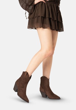 Femme portant des bottines en daim marron foncé à talons carrés et une jupe marron foncé à volants, debout devant un fond uni.